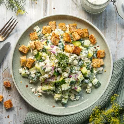 Smashed Gurkensalat mit cremigem Feta Dill Dressing und Knoblauch Croutons Smashed Gurkensalat mit Feta, roten Zwiebeln, frischem Dill und knusprigen Croutons auf einem grünen Teller, daneben ein Glas Kräuterdressing.