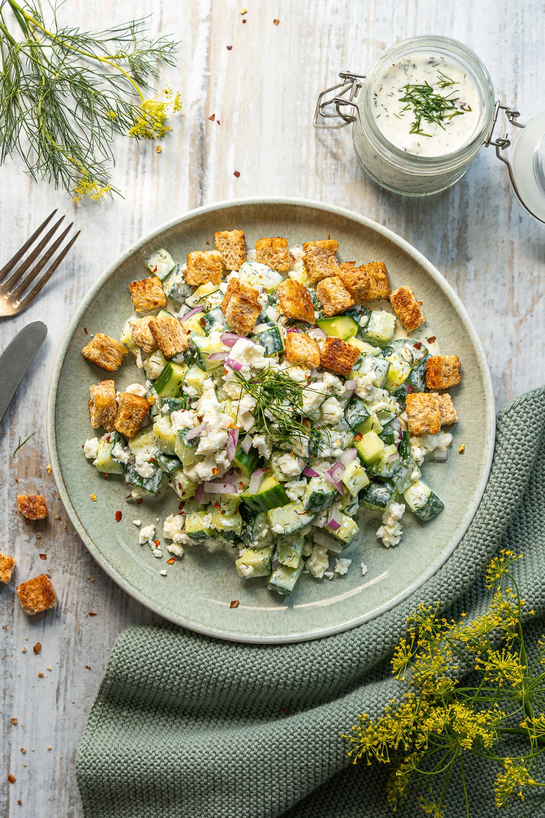 Smashed Gurkensalat mit Feta, roten Zwiebeln, frischem Dill und knusprigen Croutons auf einem grünen Teller, daneben ein Glas Kräuterdressing.