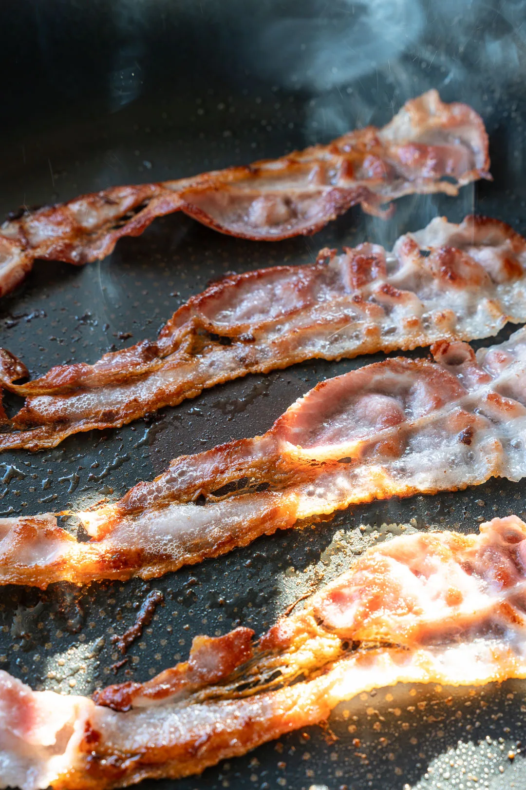 Bacon braten in einer dunklen Pfanne, mit gewellten, gebratenen Speckstreifen und Röststoffen am Boden.