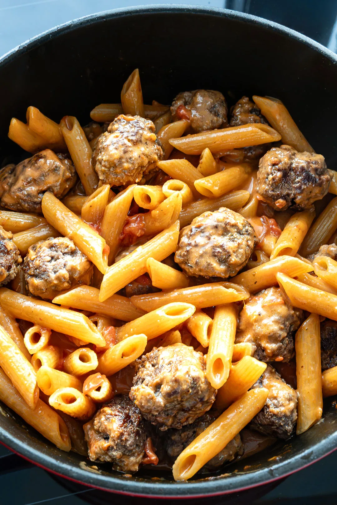 One Pot Pasta mit Meatballs in sämiger roter Sauce in einer dunklen Keramikschale, garniert mit geschmolzenem Mozzarella und frischer Petersilie.