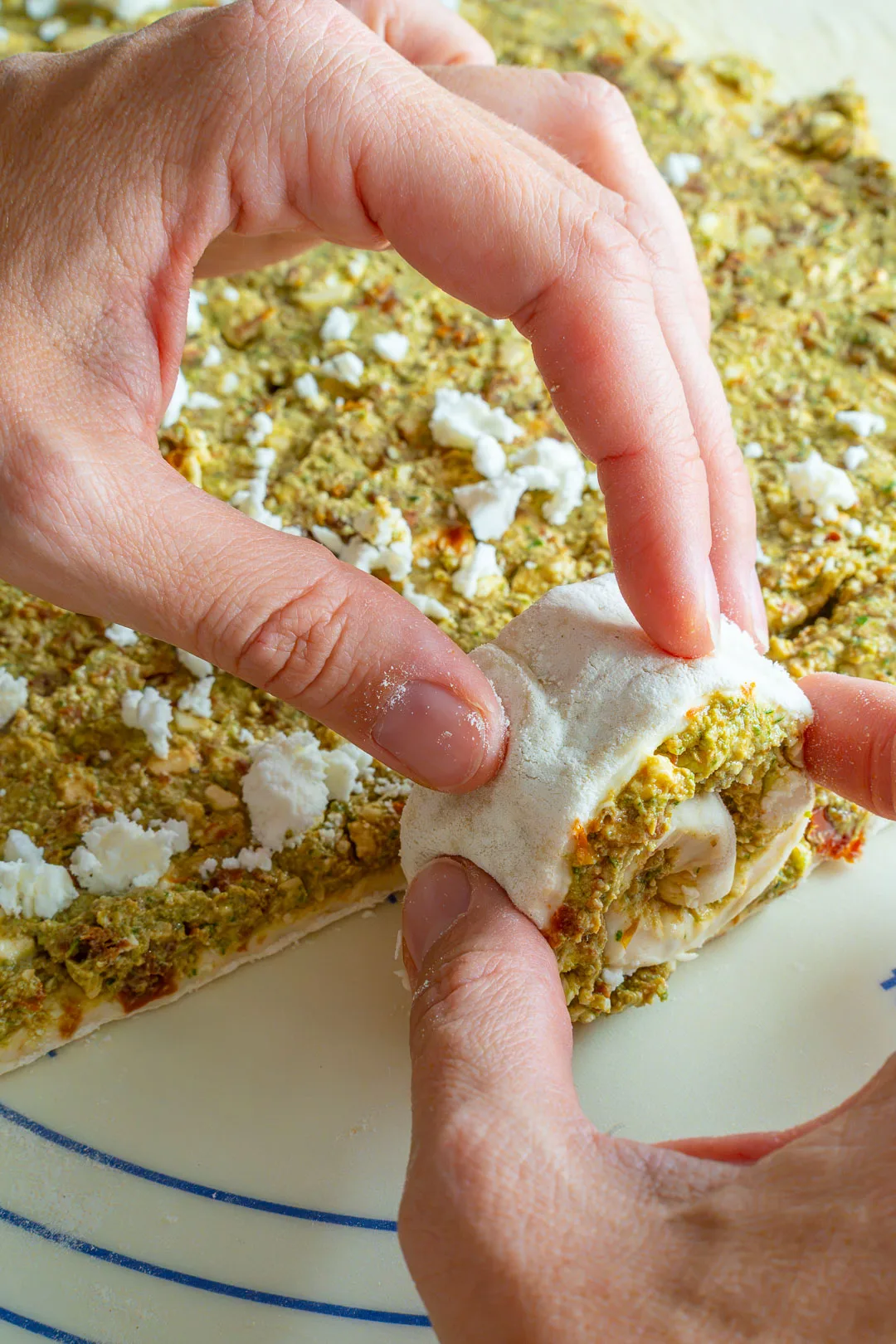 Einfaches Partygebäck: Hände rollen belegten Teig mit Pesto und Feta auf der Arbeitsfläche ein.