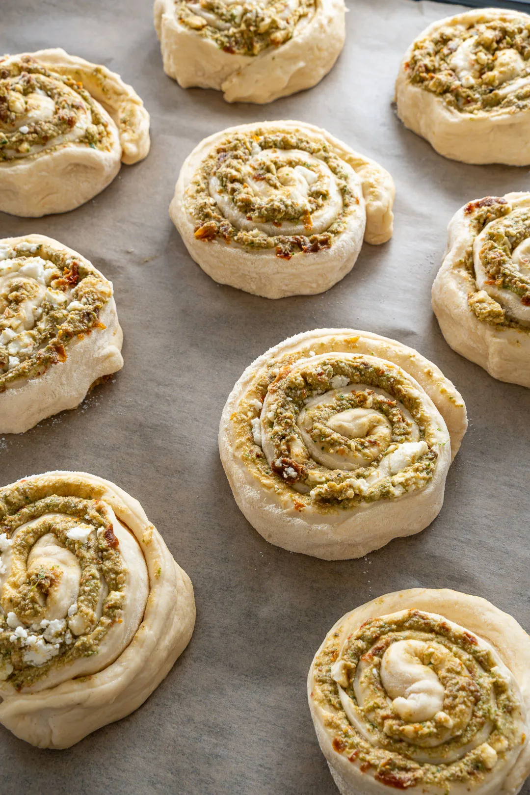 Ungebackene Pizzaschnecken mit Pesto und Feta auf einem mit Backpapier belegten Blech.