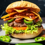 Tex Mex Burger mit Bohnen Mash auf dunklem Teller, mit Fleisch-Patty, geschmolzenem Cheddar und Zwiebelringen.