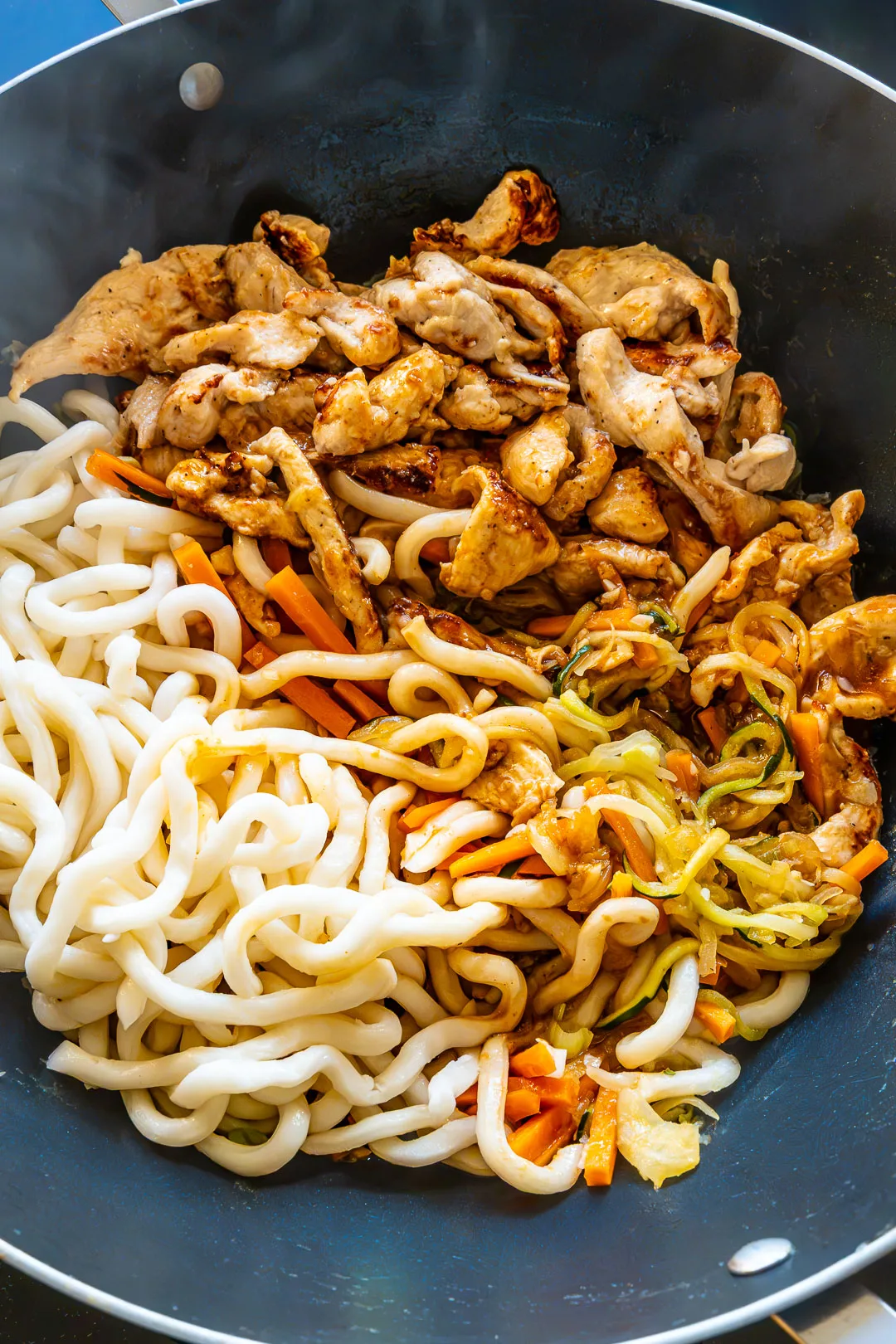 Dicke Udon-Nudeln mit Hähnchen in glänzender Sauce direkt im Wok, garniert mit Sesam und Frühlingszwiebeln.