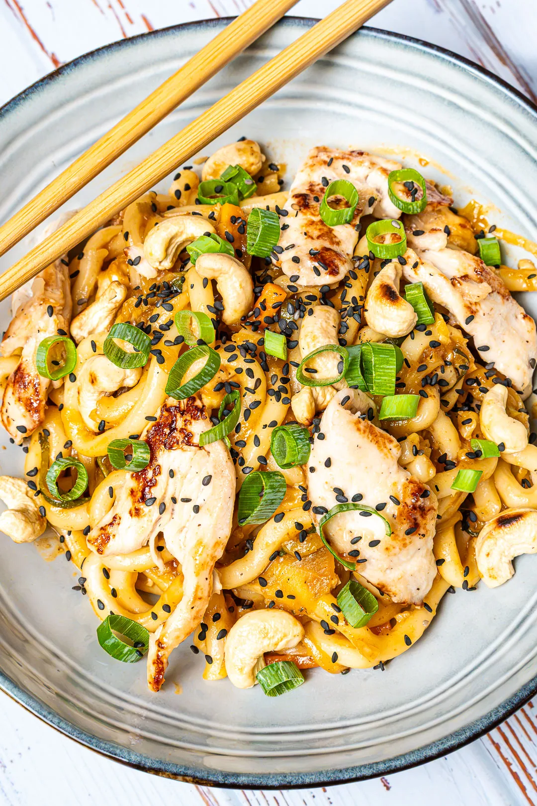 Gebratene Udon-Nudeln in glänzender Sauce, serviert in dunkler Schale mit Essstäbchen, Sesam und Frühlingszwiebeln.